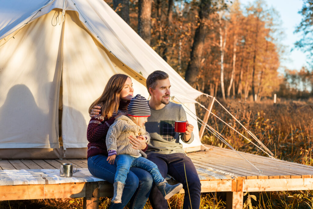 Happy family drinking coffee while sitting near canvas bell tent in the morning in a forest