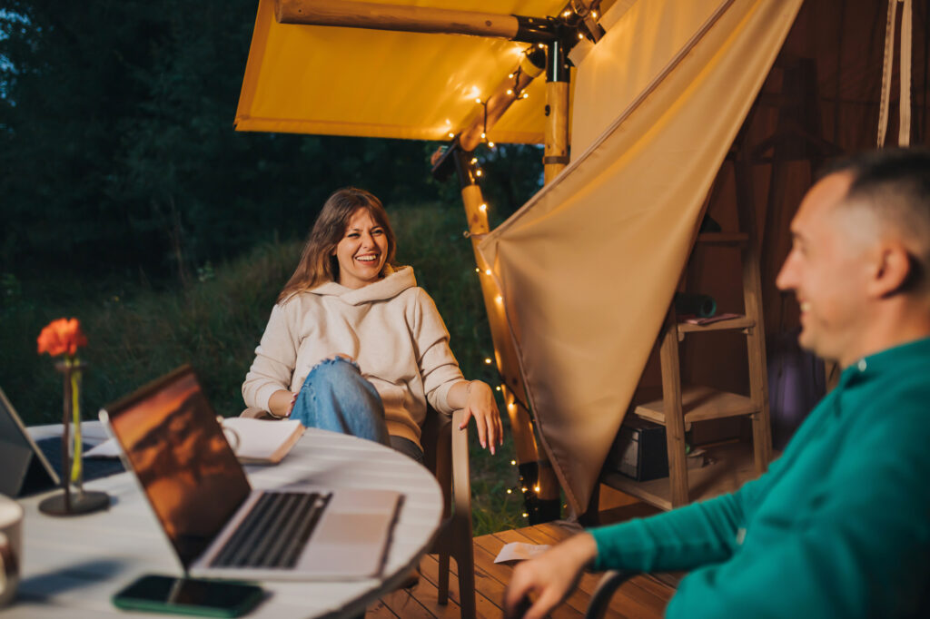 Happy family couple freelancers have a rest after working laptop on cozy glamping tent in summer evening. Luxury camping tent for outdoor holiday and vacation. Lifestyle concept